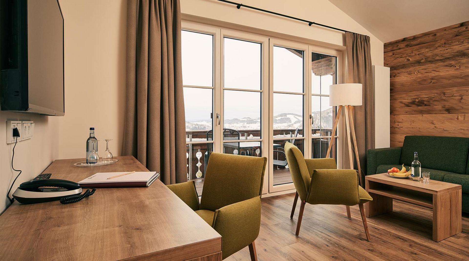  Heller Wohnbereich mit Schreibtisch, zwei grünen Sesseln, Stehlampe, Sofa und bodentiefen Fenstern mit Blick auf verschneite Landschaft.