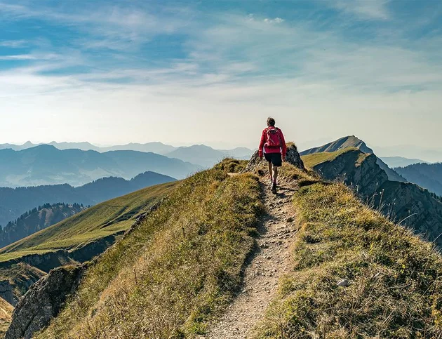 Wanderer mit rotem Rucksack auf einem schmalen Gratweg mit Bergpanorama.