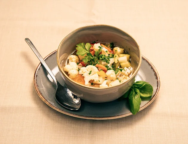 Schüssel mit Gemüsesuppe und frischen Kräutern auf einem Teller mit Löffel.