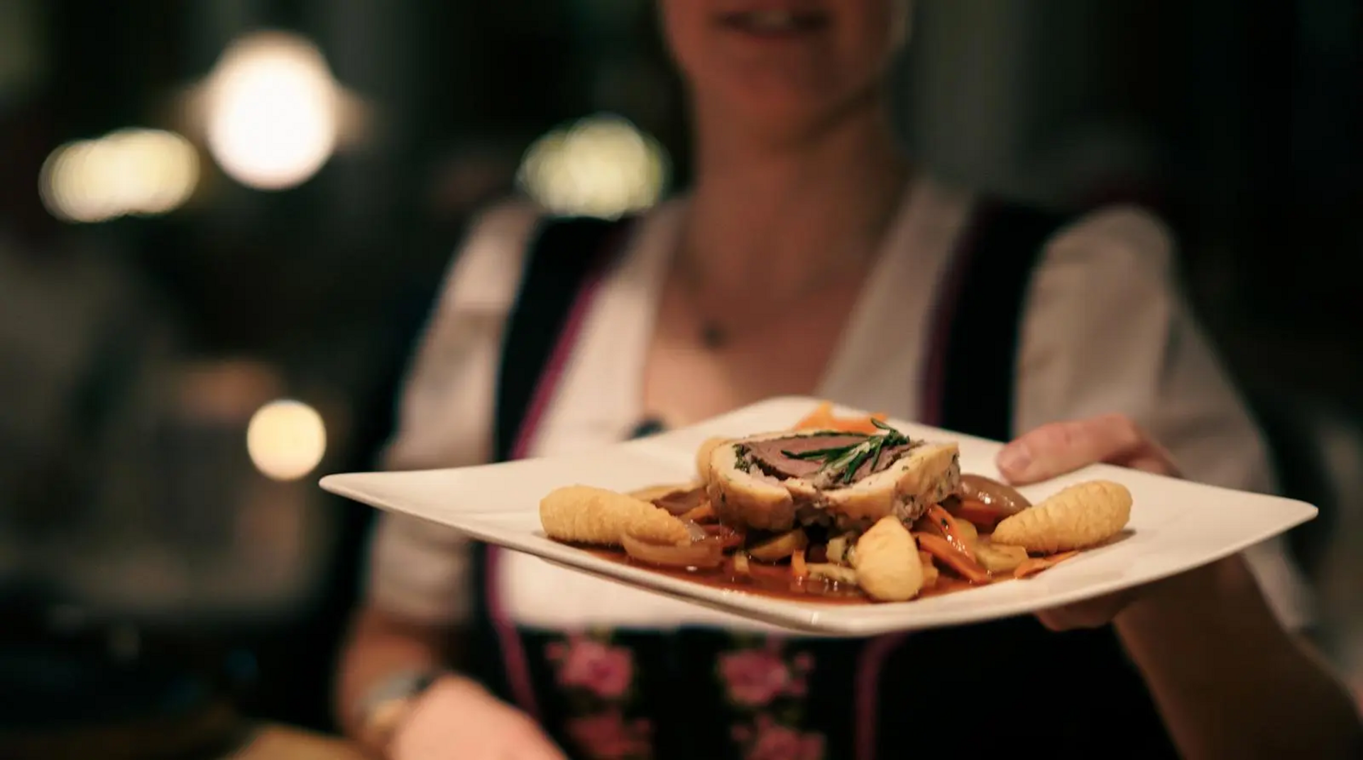 Serviererin in Tracht reicht ein stilvoll angerichtetes Gericht mit Schupfnudeln und Fleisch im Restaurant des Hotel Birkenhof Oberstaufen.