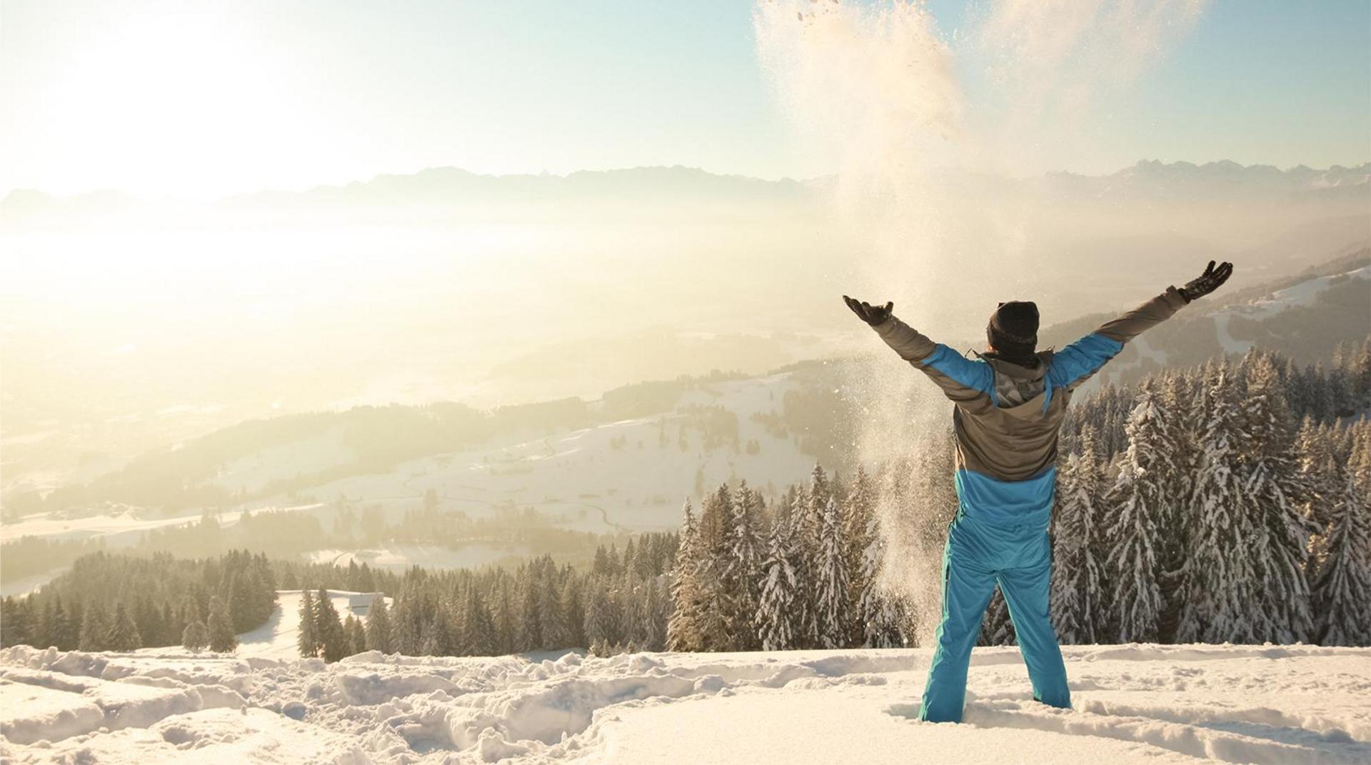 Person in Skibekleidung wirft mit ausgebreiteten Armen Schnee in die Luft, verschneite Landschaft und Berge im Hintergrund.