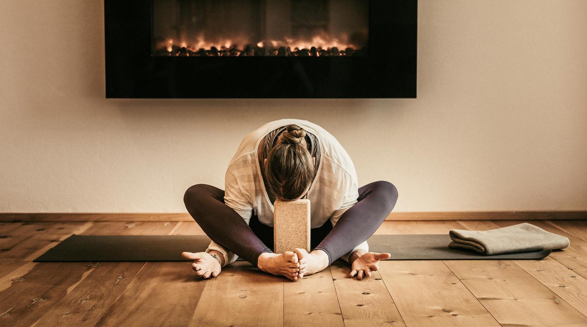 Frau sitzt auf einer Yogamatte im Schneidersitz mit nach vorne gebeugtem Oberkörper, Stirn auf einem Yogablock, im Hintergrund ein Kaminfeuer.