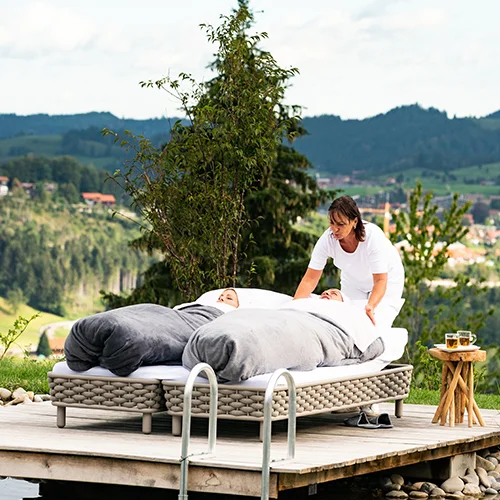 Ein Paar liegt in Schrothpackung auf Liegen im Freien beim Naturschwimmteich des Hotel Birkenhof in Oberstaufen im Allgäu, umgeben von Natur.