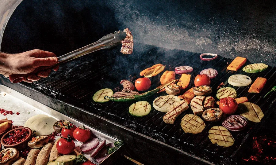 Buntes Grillgemüse und Fleischstücke brutzeln auf einem Holzkohlegrill, daneben liegen weitere frische Zutaten bereit.
