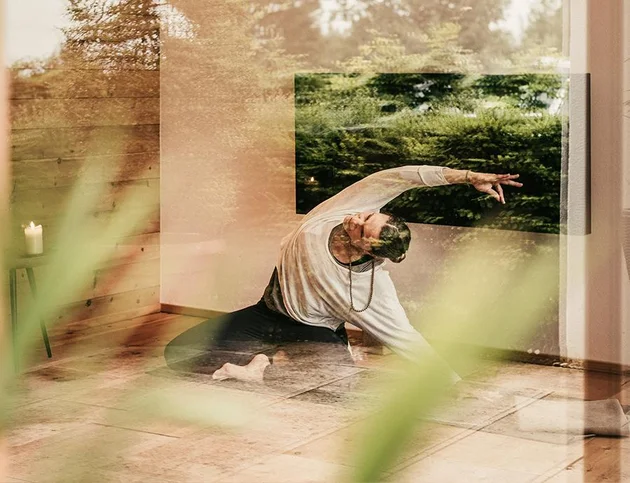 Frau beim Yoga in Dehnpose, durch Glaswand mit Naturreflexion fotografiert.