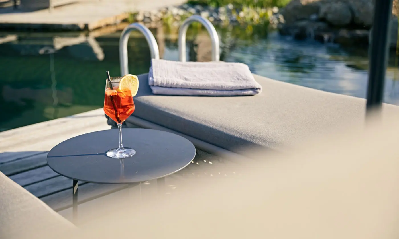 Nahaufnahme eines erfrischenden Cocktails mit Zitronenscheibe auf einem Tisch neben einer Liege am Naturteich