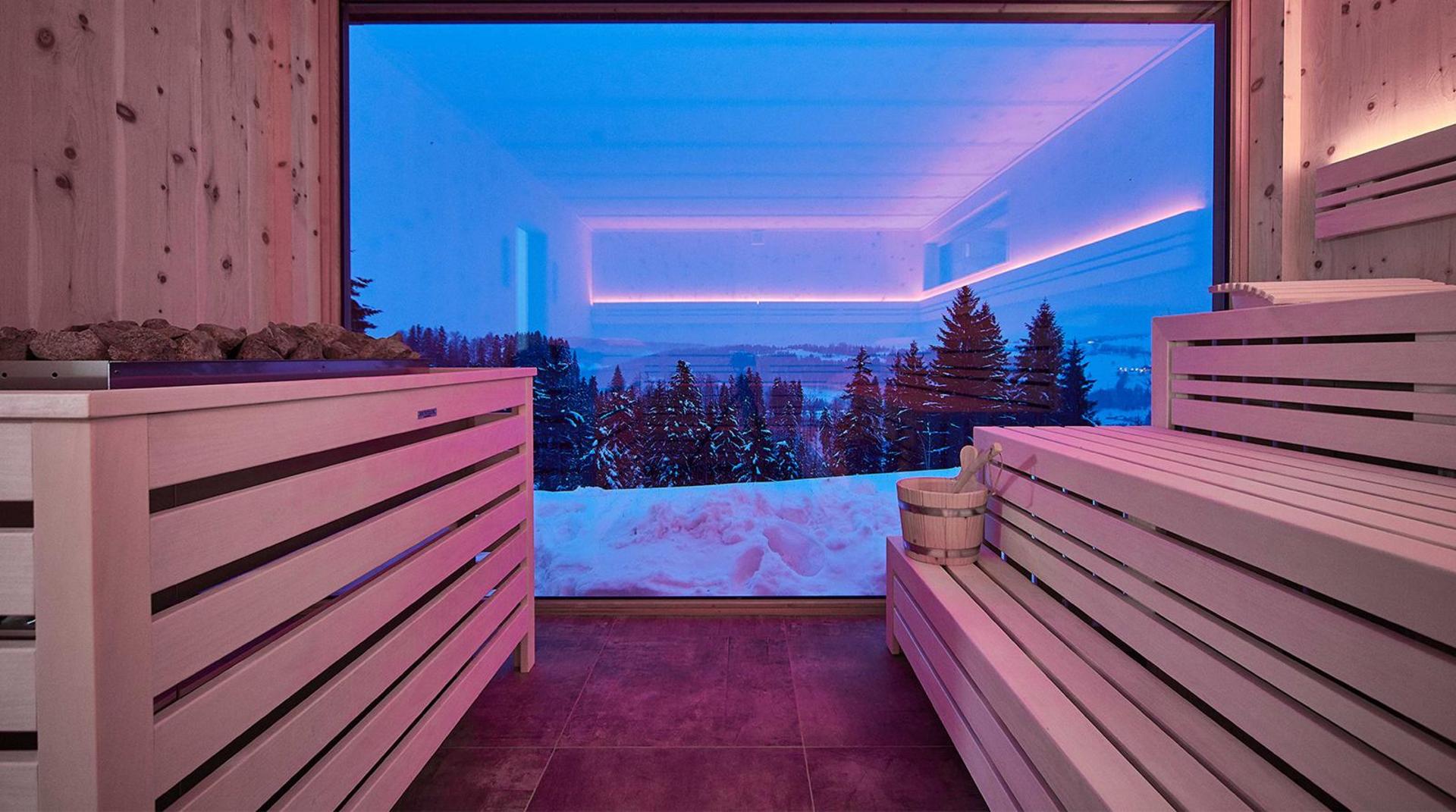 Moderne Sauna mit Holzliegen und großem Panoramafenster, Blick auf verschneite Wälder und Berge bei Abendlicht.