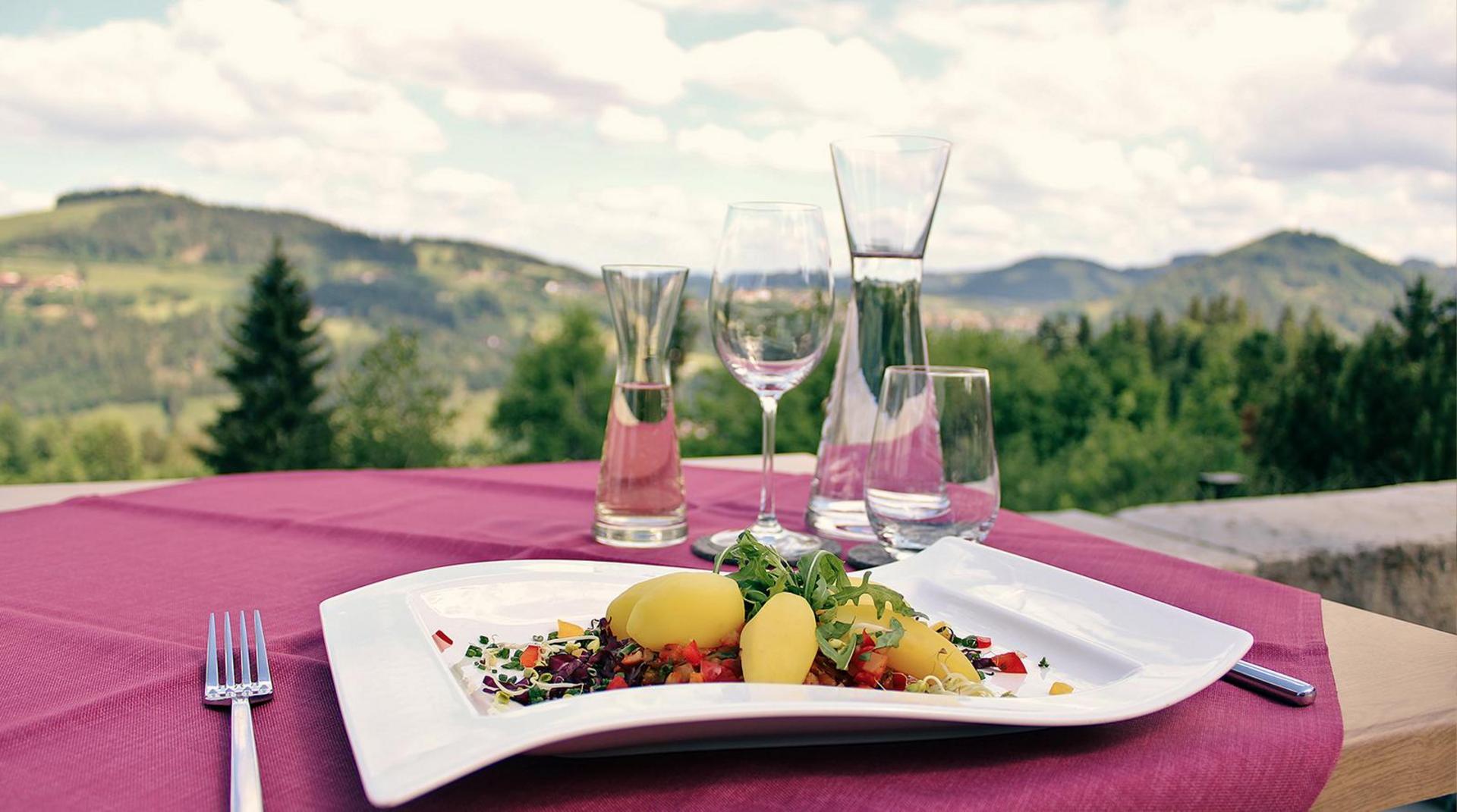 Gedeckter Tisch auf Terrasse mit Bergpanorama, Glasflaschen und elegant angerichtetem Tellergericht.