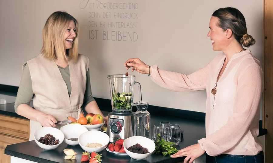 Zwei Frauen bereiten gemeinsam grüne Smoothies mit frischem Obst und Gemüse zu.