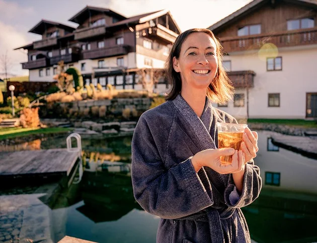 Frau im Bademantel lächelt und hält ein Getränk in der Hand, im Hintergrund der Naturschwimmteich und das Hotel Birkenhof in Oberstaufen.