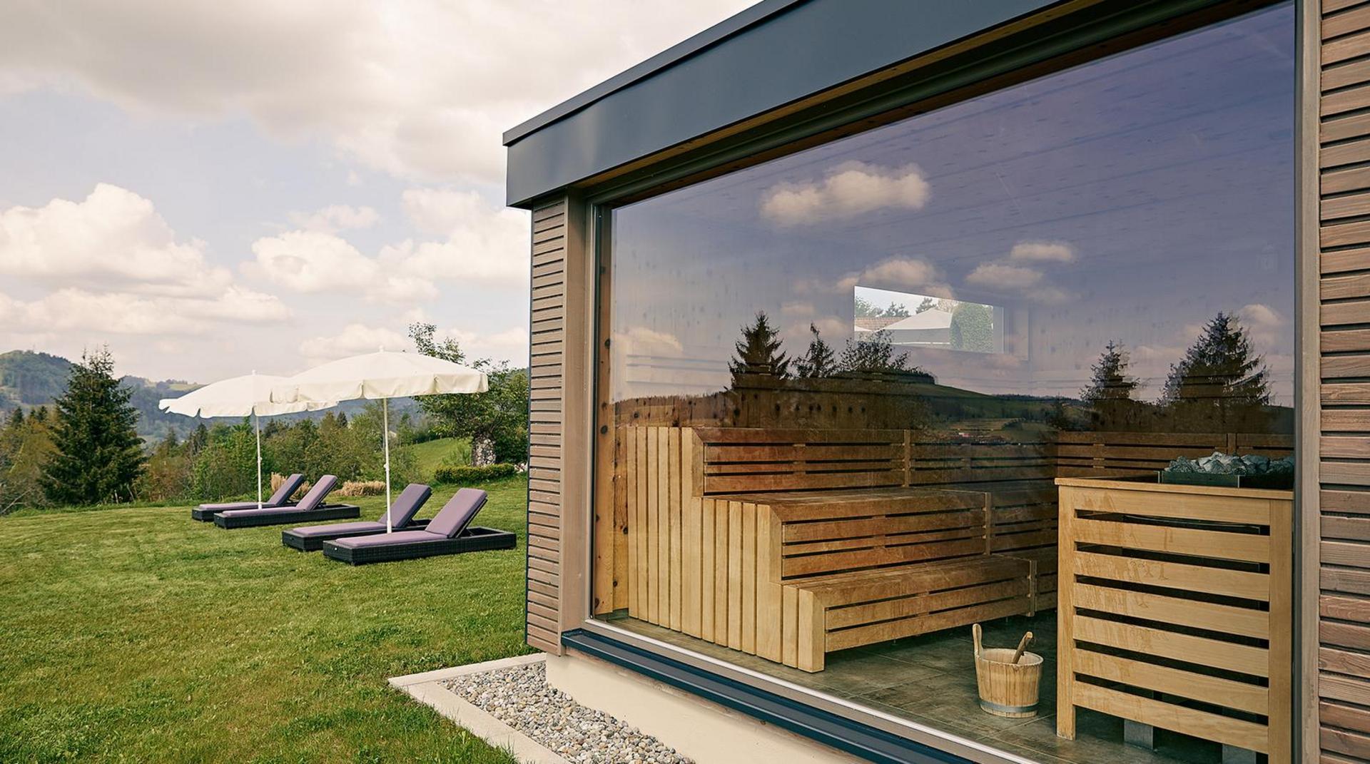 Moderne Sauna mit Panoramafenster im Hotel Birkenhof und Blick auf den Garten mit Sonnenliegen und Schirmen.
