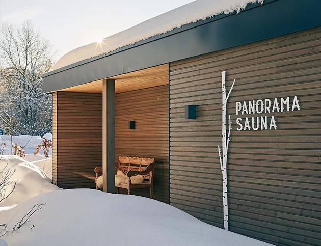 Eingangsbereich der Panorama-Sauna im Winter, moderne Holzfassade mit Beschriftung ‚Panorama Sauna‘, davor Bank mit Fellen, umgeben von tiefem Schnee.