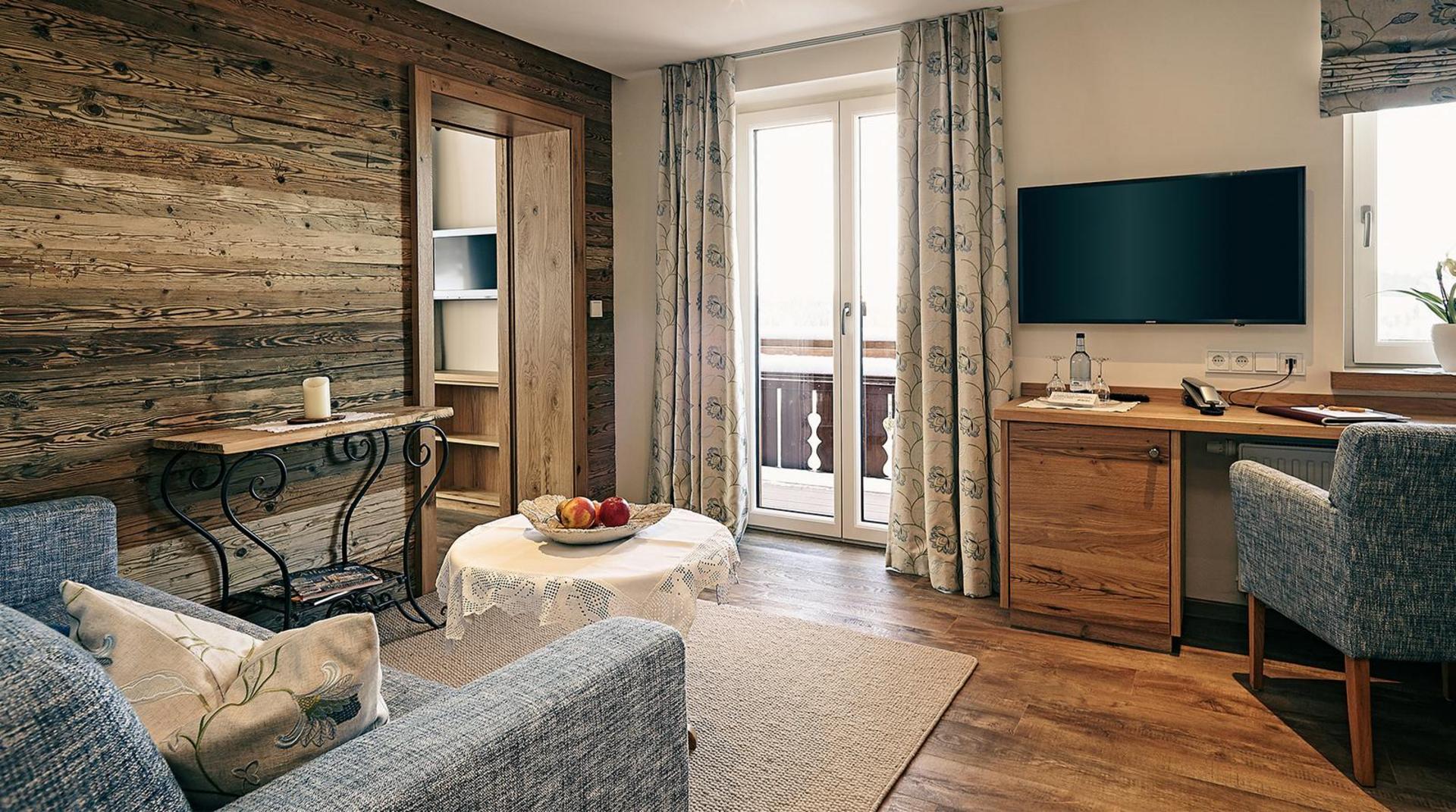 Gemütlicher Wohnbereich mit Holzwand, grauen Polstersesseln, rundem Tisch mit Obstschale, Schreibtisch, Fernseher und Zugang zum Balkon.