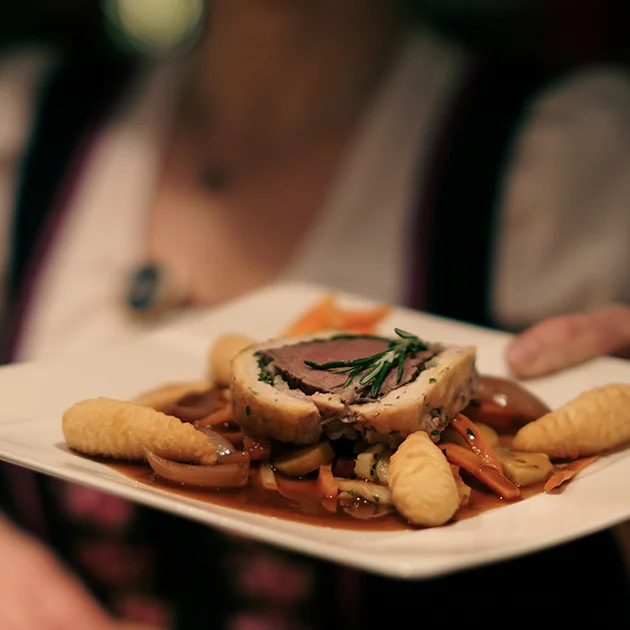 Eine Kellnerin serviert ein traditionelles Fleischgericht mit Kroketten, angerichtet auf einem eleganten weißen Teller