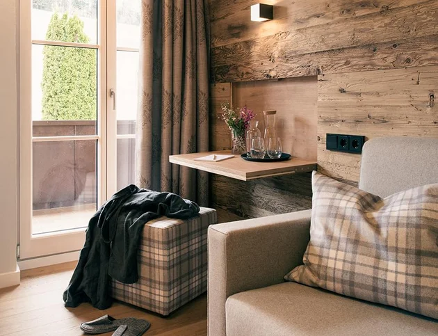Helles Einzelzimmer mit Sofa, kariertem Hocker und kleinem Wandtisch vor Holzwand, daneben Balkontür mit Vorhängen.