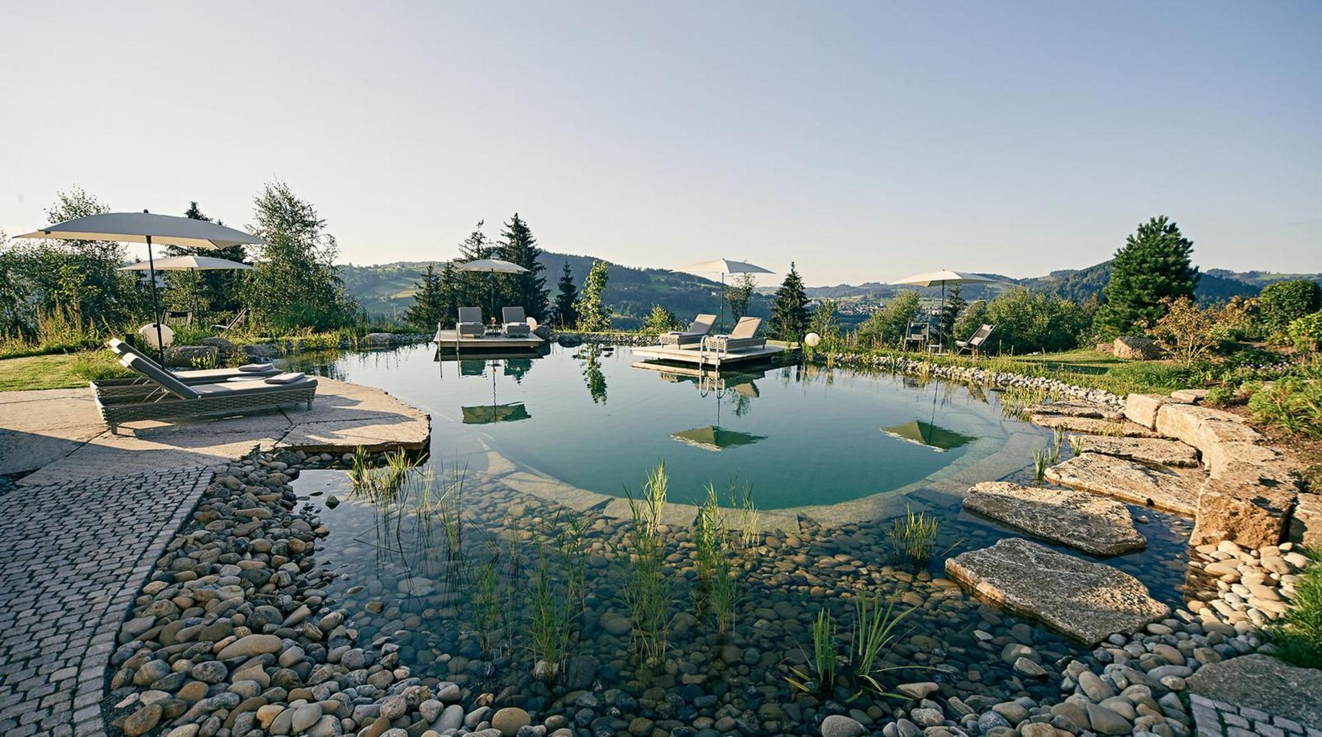 Naturschwimmteich im Sommer mit Sonnenliegen, Sonnenschirmen und Blick auf die Allgäuer Landschaft beim Hotel Birkenhof.