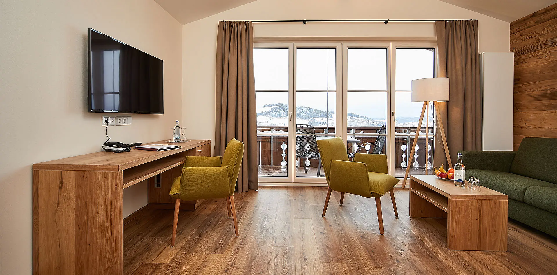Helles Wohnzimmer mit zwei grünen Sesseln, Sofa, Schreibtisch und großem Fenster mit Bergblick