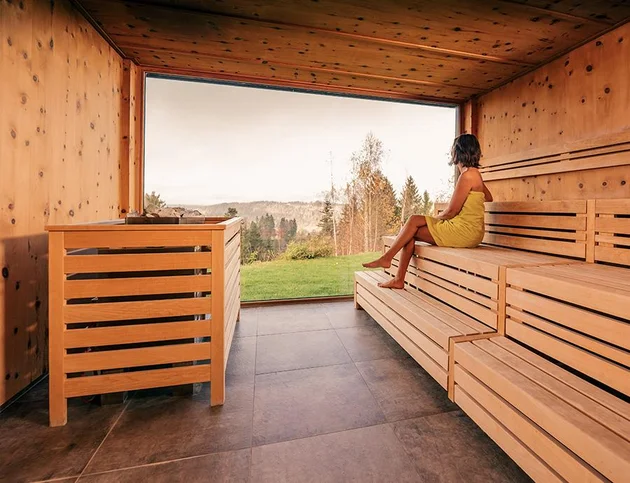 Frau sitzt in einer Holzsauna auf den Bänken und blickt durch ein großes Panoramafenster auf Wiesen und Bäume.
