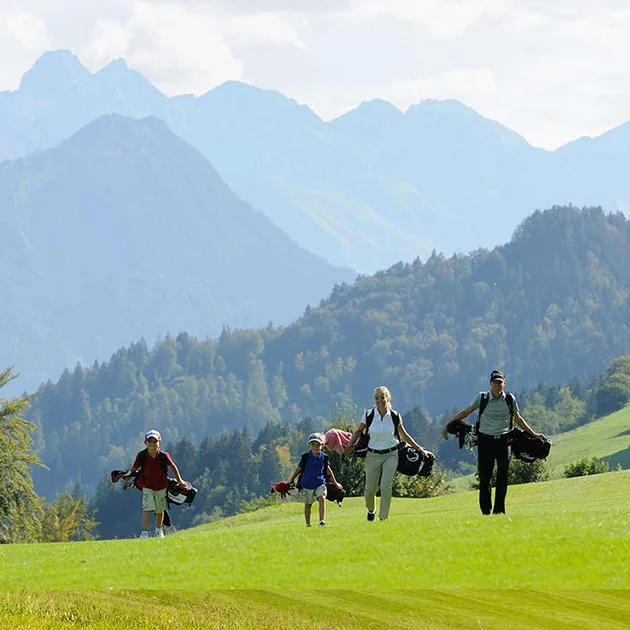 Familie mit zwei Kindern trägt Golfausrüstung über einen sonnigen Golfplatz mit Bergkulisse.