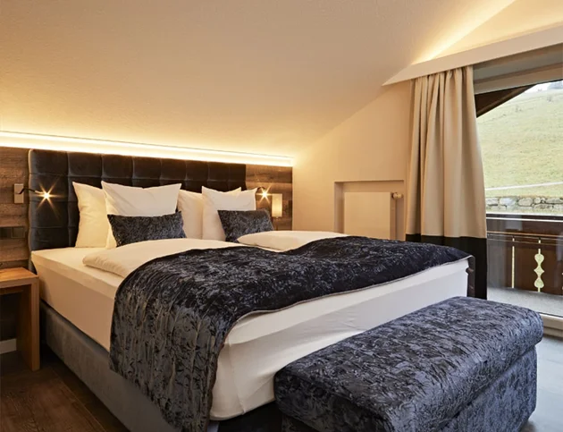  Doppelzimmer mit Dachschräge, gepolstertem Kopfteil, dunkelblauer Bettwäsche, Sitzbank am Fußende und Zugang zum Balkon.