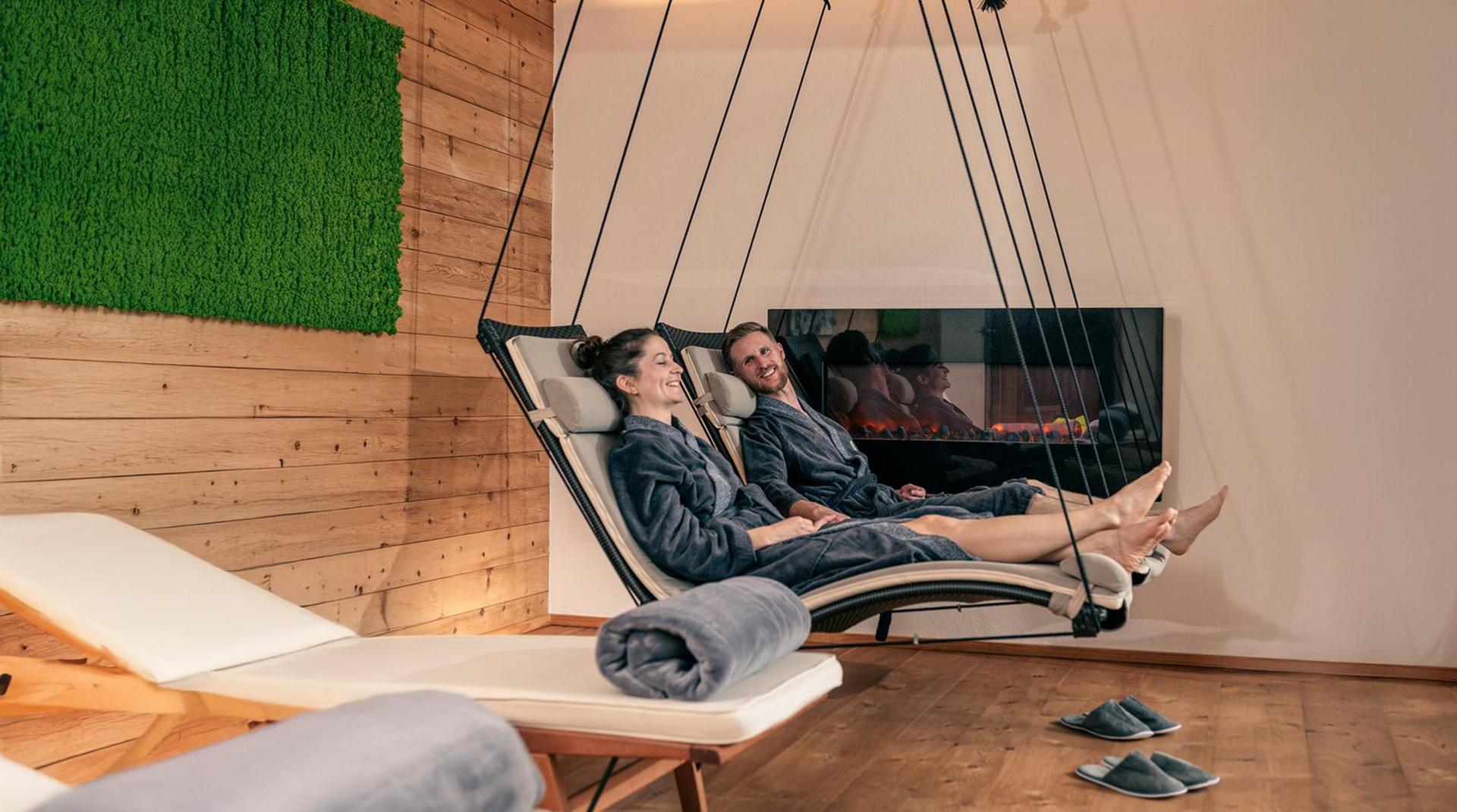 Paar entspannt in schwebenden Relaxliegen im Raum der Stille des NaturSpa im Hotel Birkenhof Oberstaufen, vor einem Kaminfeuer.