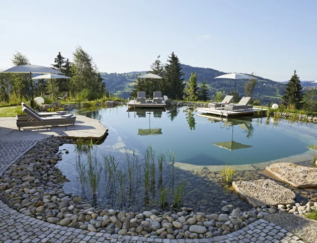 Natürlicher Schwimmteich mit Liegen und Sonnenschirmen auf Holzplattformen, umgeben von Steinen, Pflanzen und Blick auf die Berge.