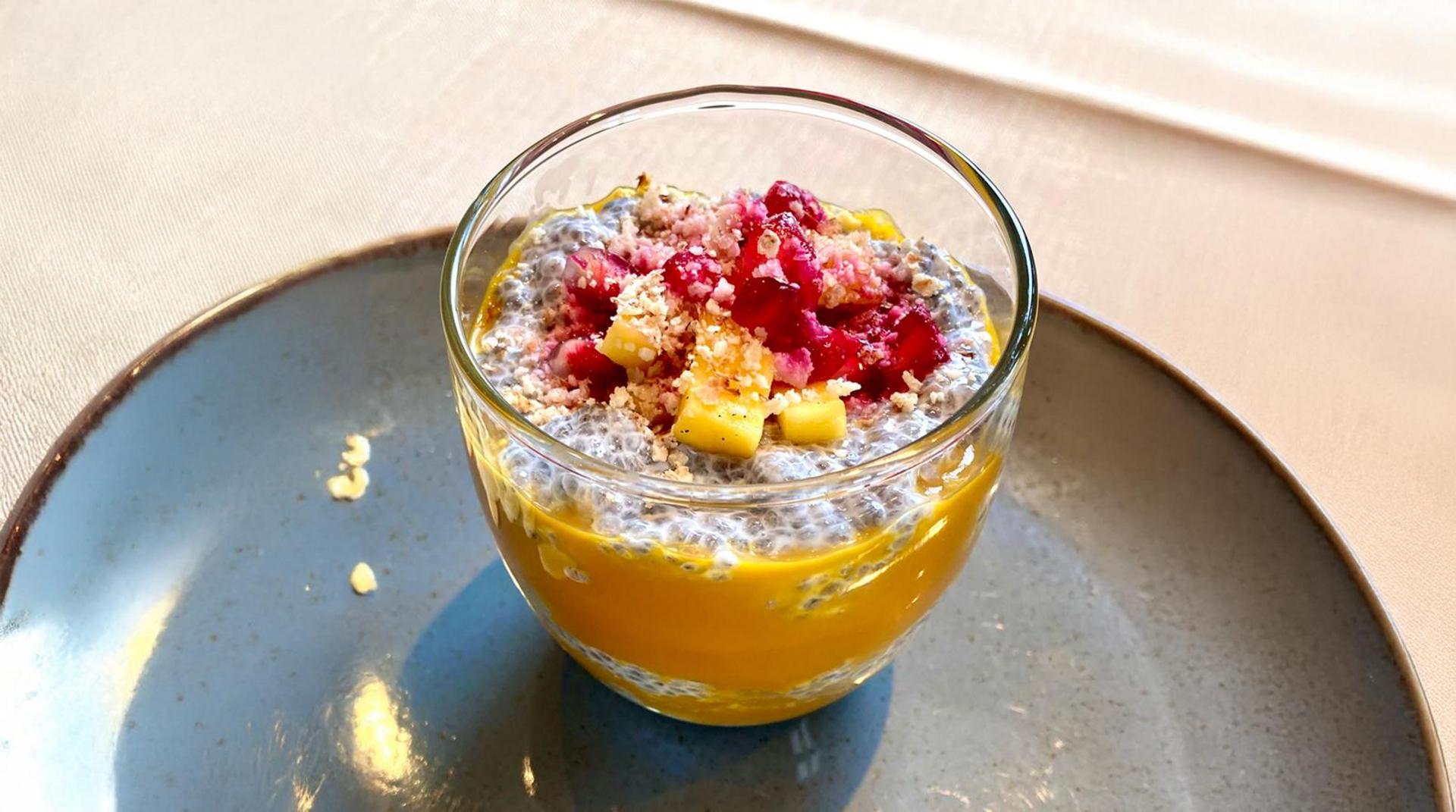 Dessert im Glas mit gelbem Fruchtpüree, Chiasamen-Schicht, Himbeeren und Kokosraspeln auf einem blauen Teller.