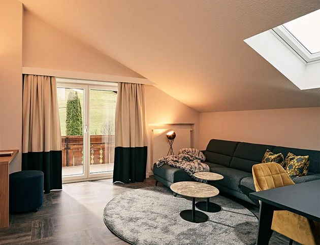 Helles Wohnzimmer unter Dachschräge mit grauem Ecksofa, gemusterten Kissen, zwei runden Beistelltischen, gelbem Stuhl und Zugang zum Balkon.