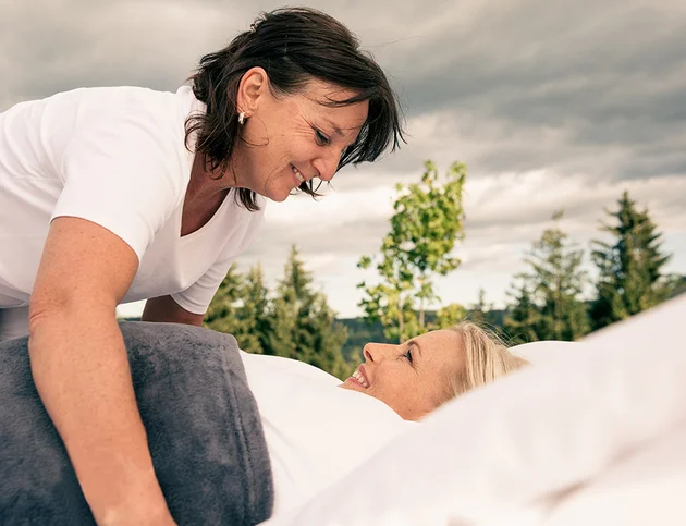 Frau beugt sich lächelnd über eine andere Frau, die eingekuschelt im Bett liegt.
