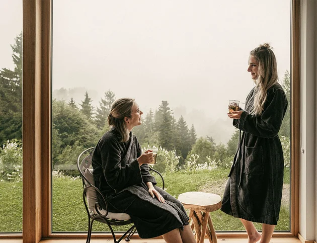 Zwei Frauen im Bademantel sitzen an einem Fenster mit Blick ins Grüne und trinken Tee.