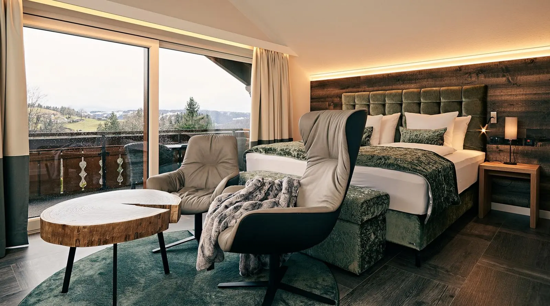  Gemütliches Hotelzimmer mit Doppelbett, zwei Sesseln und rustikalem Holztisch vor bodentiefem Fenster mit Blick auf einen Balkon und grüne Hügellandschaft.
