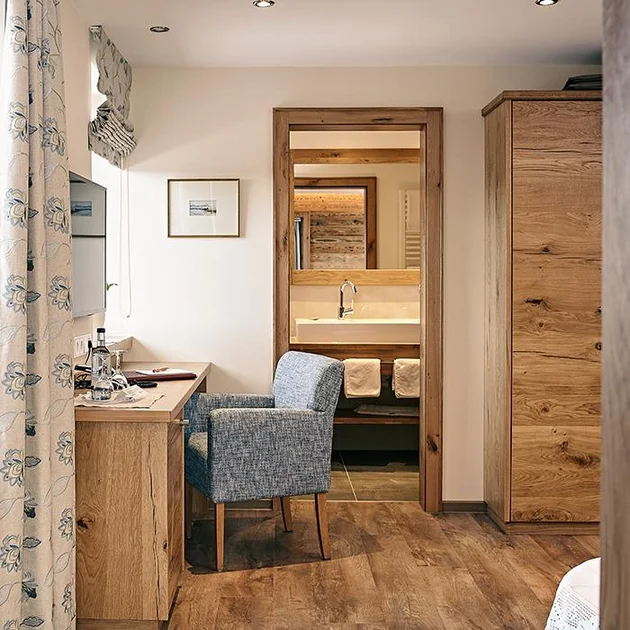 Gemütliches Hotelzimmer im Birkenhof Oberstaufen mit Holzschreibtisch, blauem Sessel und Blick ins moderne Badezimmer.