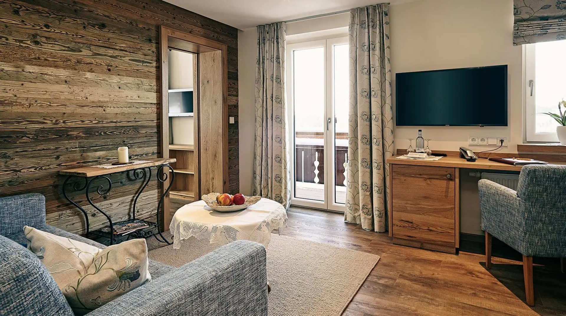 Gemütlicher Wohnbereich mit Holzwand, grauen Polstersesseln, rundem Tisch mit Obstschale, Schreibtisch, Fernseher und Zugang zum Balkon.