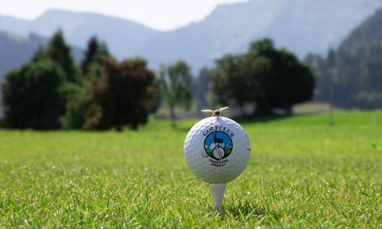 Golfball mit Logo des Golfclubs Oberstaufen-Steibis auf Tee, im Hintergrund grüne Fairways und Berge
