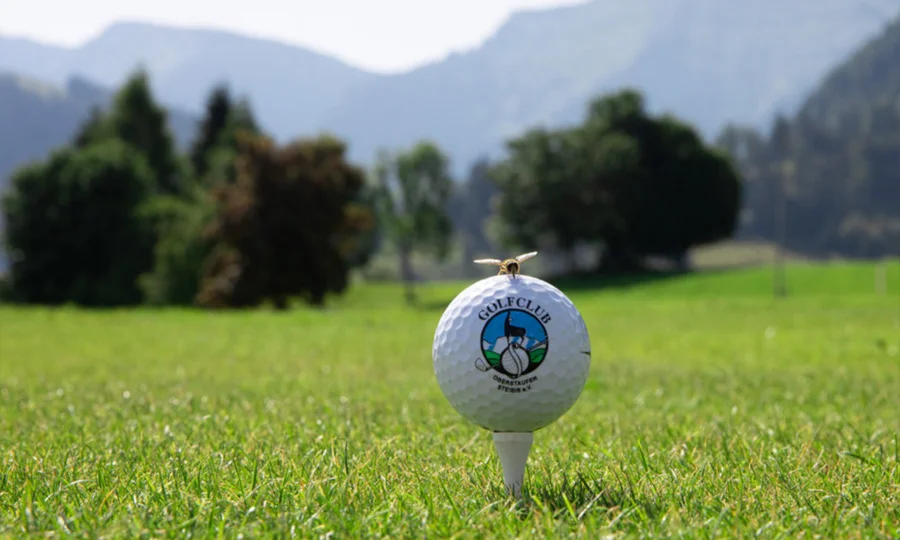Golfball mit Logo des Golfclubs auf Tee, dahinter Wiese und Berge.