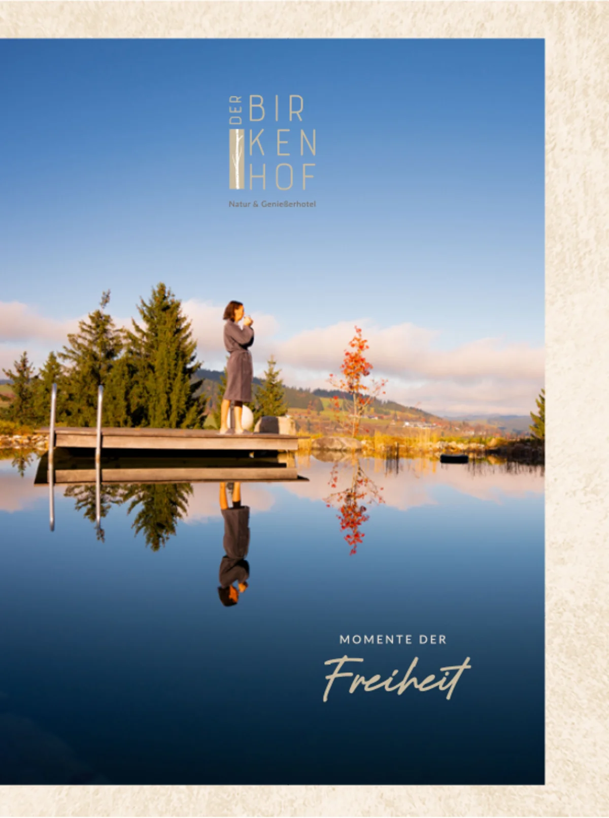 Titelseite der Broschüre „Der Birkenhof – Natur & Genusshotel“: Frau steht auf einem Holzsteg am ruhigen See, klare Spiegelung im Wasser, dahinter herbstliche Bäume und sanfte Hügel. Text: „Momente der Freiheit“.
