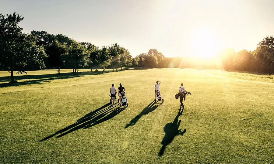Vier Golfer gehen mit Schlägertaschen über einen sonnigen Golfplatz in Richtung Sonnenuntergang.