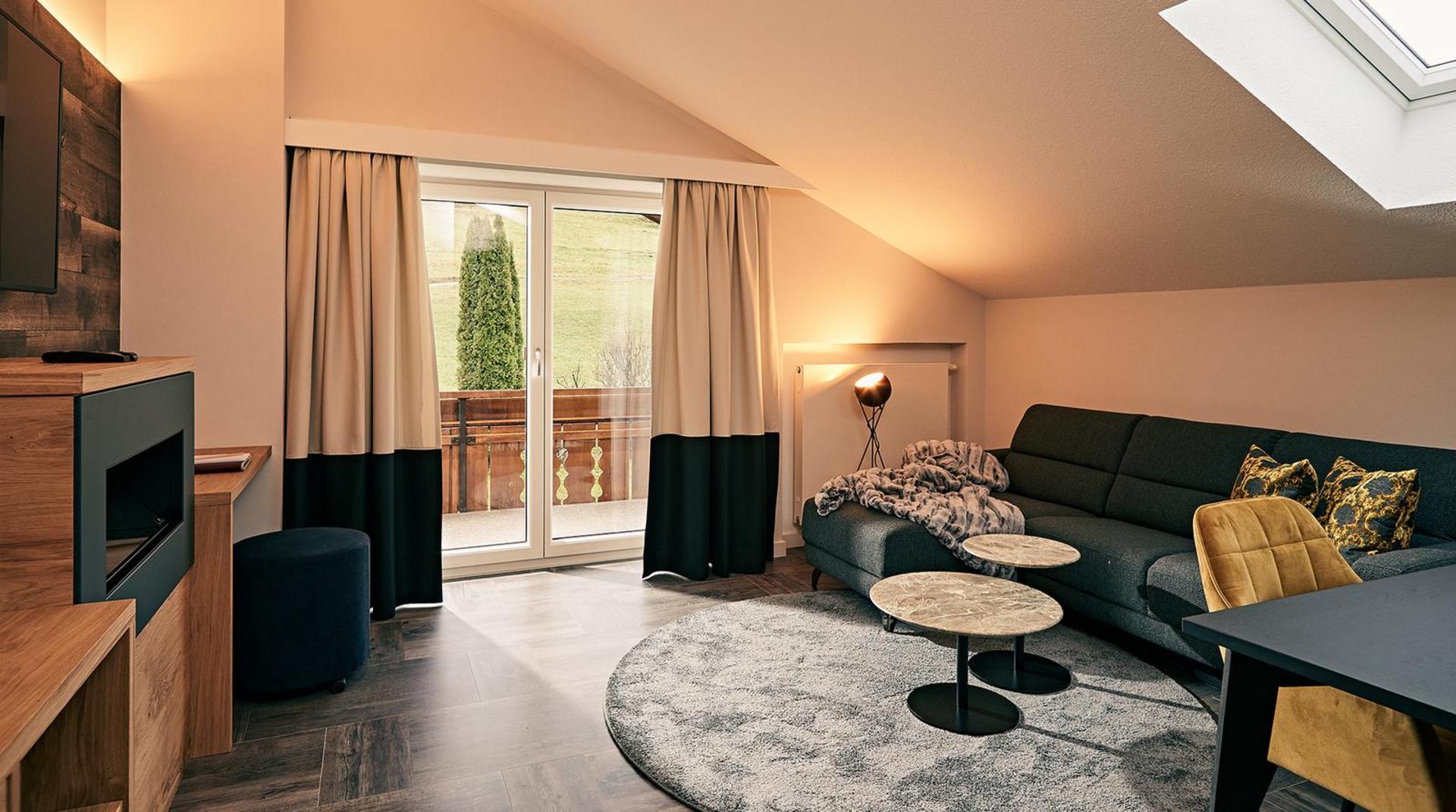  Helles Wohnzimmer mit Dachschräge, grauem Ecksofa, zwei runden Beistelltischen, gelbem Sessel und Zugang zu einem Balkon mit Blick ins Grüne.