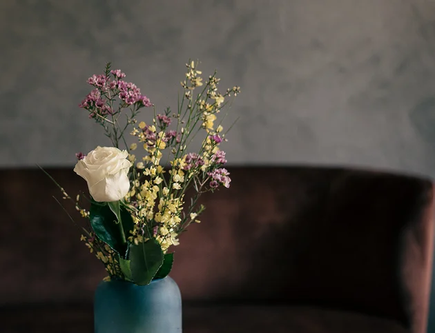 Vase mit weißer Rose und bunten Trockenblumen auf einem Tisch vor dunklem Hintergrund.