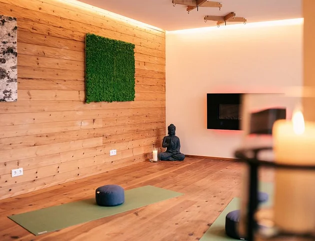 Yoga- und Meditationsraum mit Holzwand, grüner Moosdeko, Yoga-Matten, Buddha-Statue und Kerzenlicht.