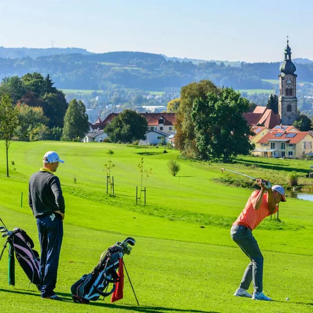 Zwei Golfer auf dem Golfplatz Schlossgut Oberstaufen, einer beim Abschlag, im Hintergrund die Kirche und Häuser des Ortes mit grüner Landschaft.