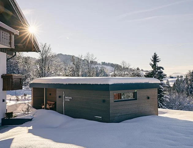 Außenansicht einer modernen Panoramasauna im Winter, umgeben von tiefem Schnee, mit Holzverkleidung und kleinem Fenster; im Hintergrund verschneite Bäume und sanfte Hügel bei Sonnenschein.