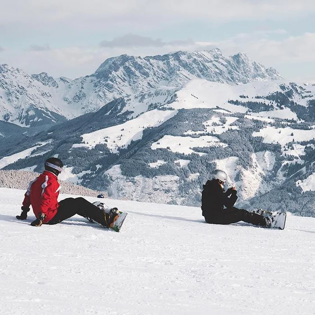 Zwei Snowboarder sitzen im Schnee mit Blick auf die verschneiten Berge am Hochgrat bei Oberstaufen im Allgäu.