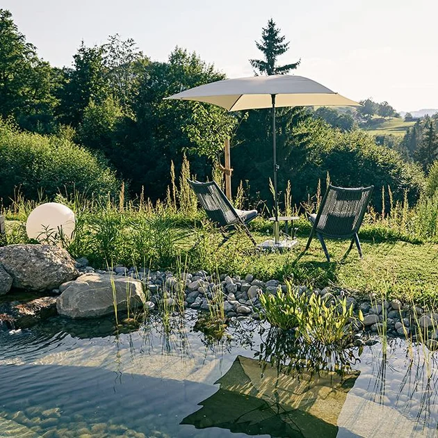 Naturschwimmteich mit Blick in das Grüne zwei Gartenliegen und ein Sonnenschirm