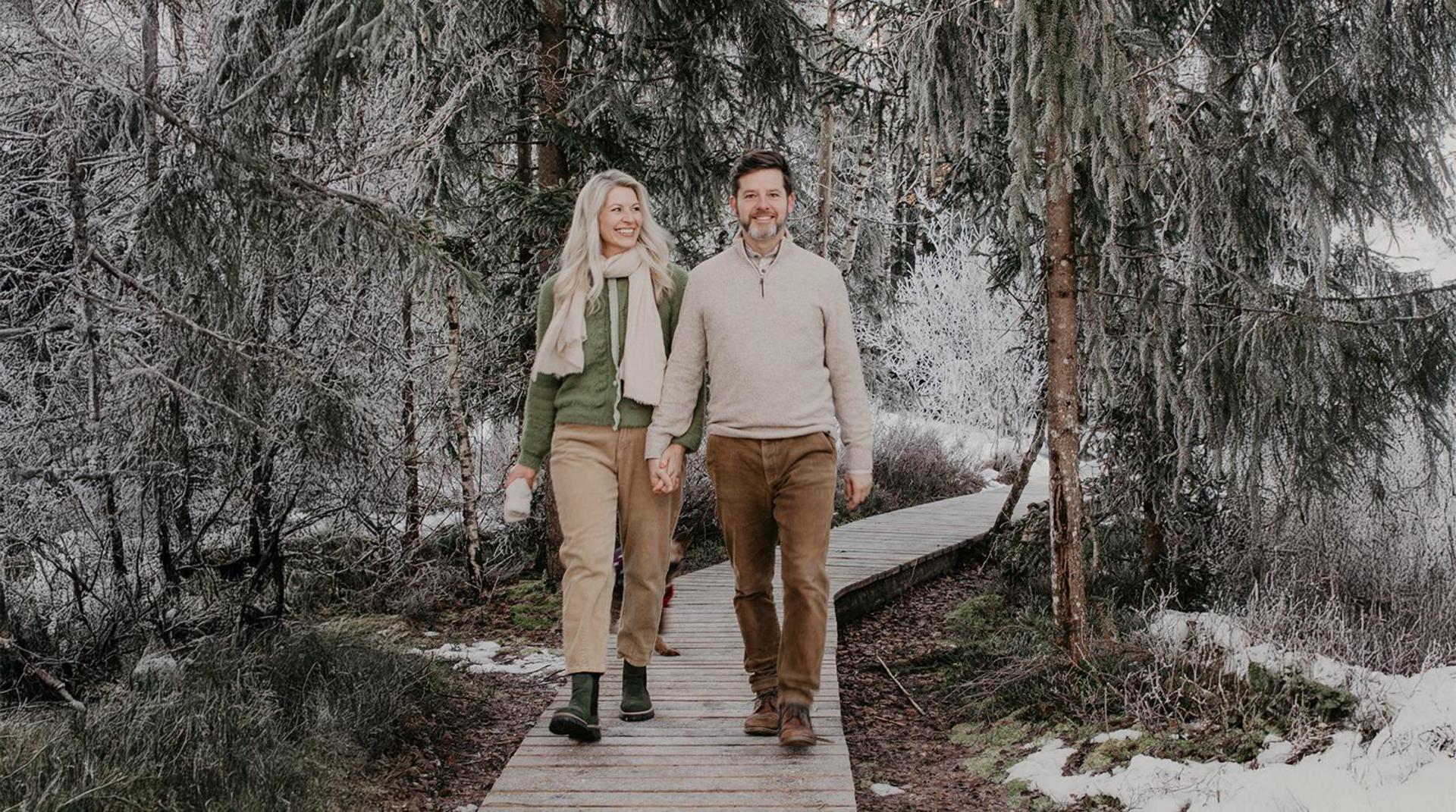 Andrea und Thomas spazieren Hand in Hand auf Holzsteg durch verschneiten Winterwald nahe dem Hotel Birkenhof in Oberstaufen
