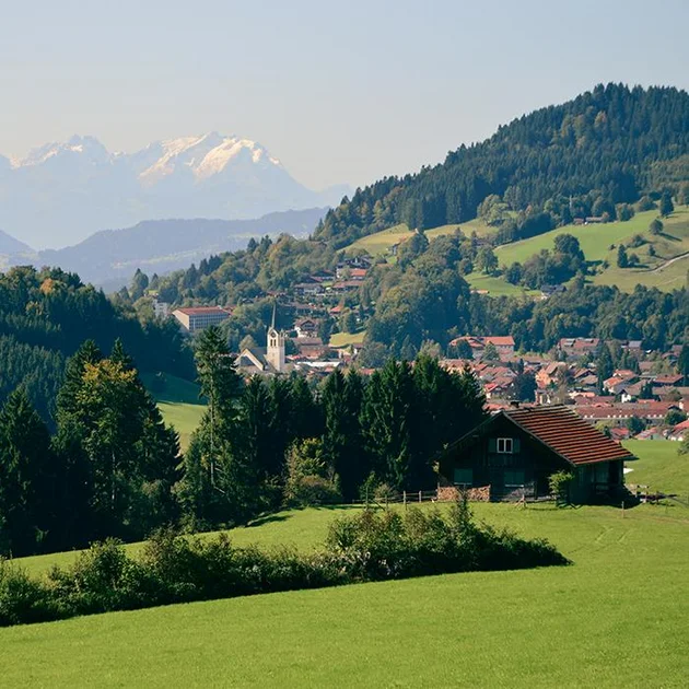 Blick auf Oberstaufen im Sommer mit grünen Wiesen, Wäldern, der Kirche im Ort und den Alpen im Hintergrund.