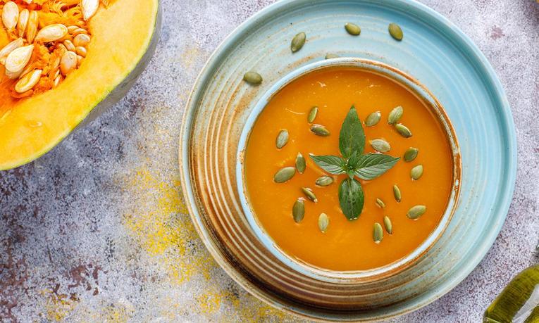 Teller mit Kürbissuppe, garniert mit Kürbiskernen und frischen Kräutern, daneben aufgeschnittener Kürbis