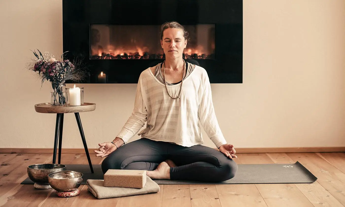 Frau sitzt im Schneidersitz auf einer Yogamatte vor einem Kamin und meditiert