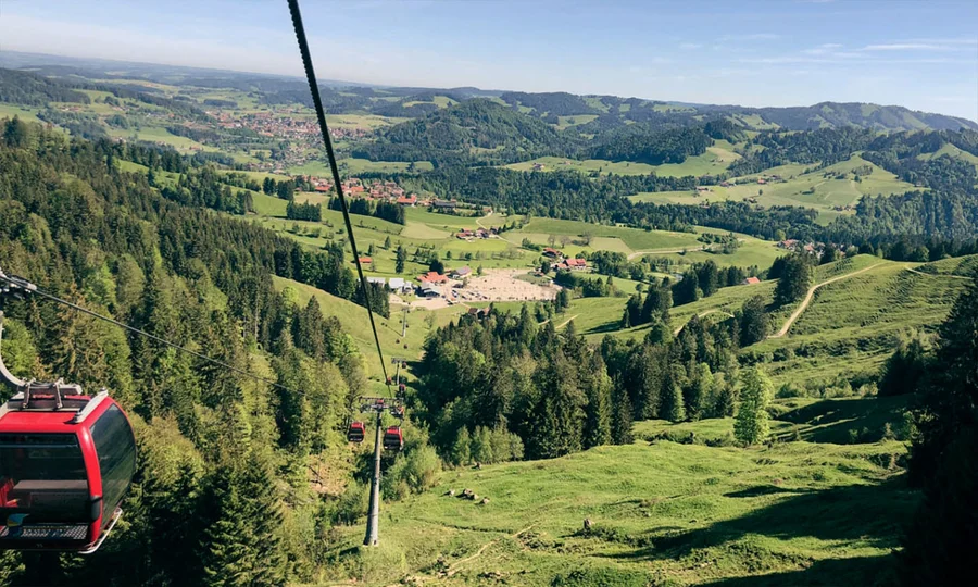 Rote Gondelbahn fährt über grüne Hügel und Wälder hinab ins Tal mit verstreuten Häusern und weitem Blick über die Allgäuer Landschaft.