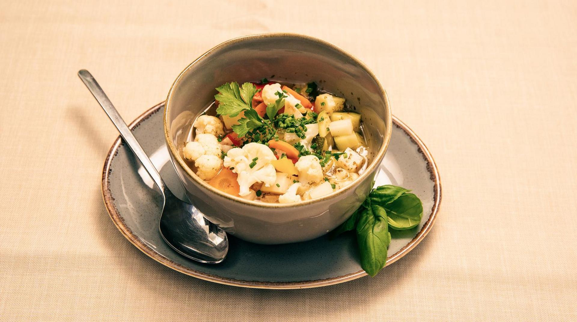 Schüssel mit Gemüsesuppe auf einem Teller, daneben ein Löffel und frisches Basilikum.
